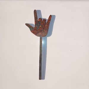 May include: A rusty metal sculpture of a hand making the sign for 'I love you' in American Sign Language. The hand is attached to a long, thin metal rod.