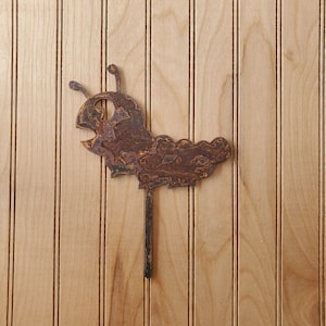May include: A rusty metal garden stake shaped like a cartoon caterpillar with a large eye and a long, thin body. The stake is attached to a wooden surface.