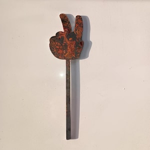 May include: A rusty metal peace sign with a long stem. The peace sign is a hand with two fingers raised in the peace sign gesture.