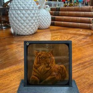 May include: A square wooden coaster with a black frame and base, displaying a detailed image of a tiger. The word "TIGER" is printed at the top. The coaster is on a wooden surface, with decorative pineapples and books in the background.