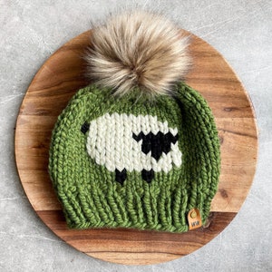 May include: Green knitted hat with a white sheep design and a brown faux fur pom pom. The hat has a brown leather tag with the letters "WW" on it.