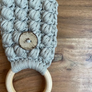 May include: A light gray macrame towel holder featuring a textured bobble stitch pattern, a single large round wooden ring at the base, and a circular wooden button centered on the front of the woven fabric construction.