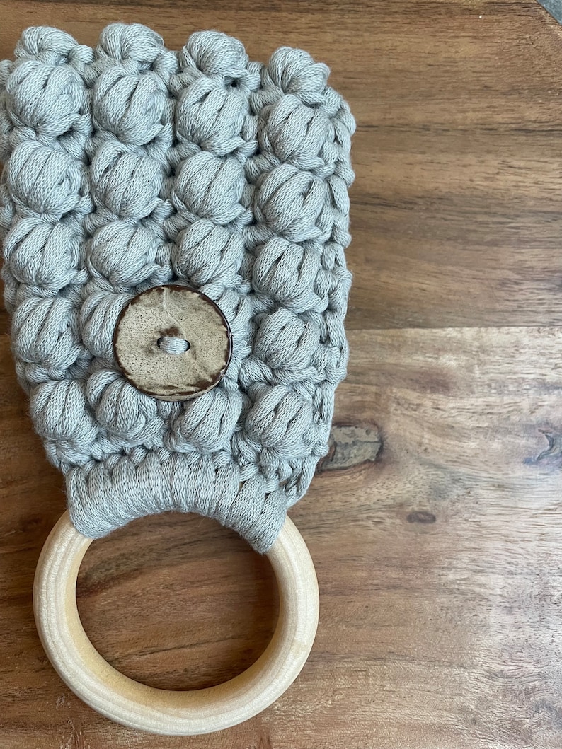 May include: A light gray macrame towel holder featuring a textured bobble stitch pattern, a single large round wooden ring at the base, and a circular wooden button centered on the front of the woven fabric construction.