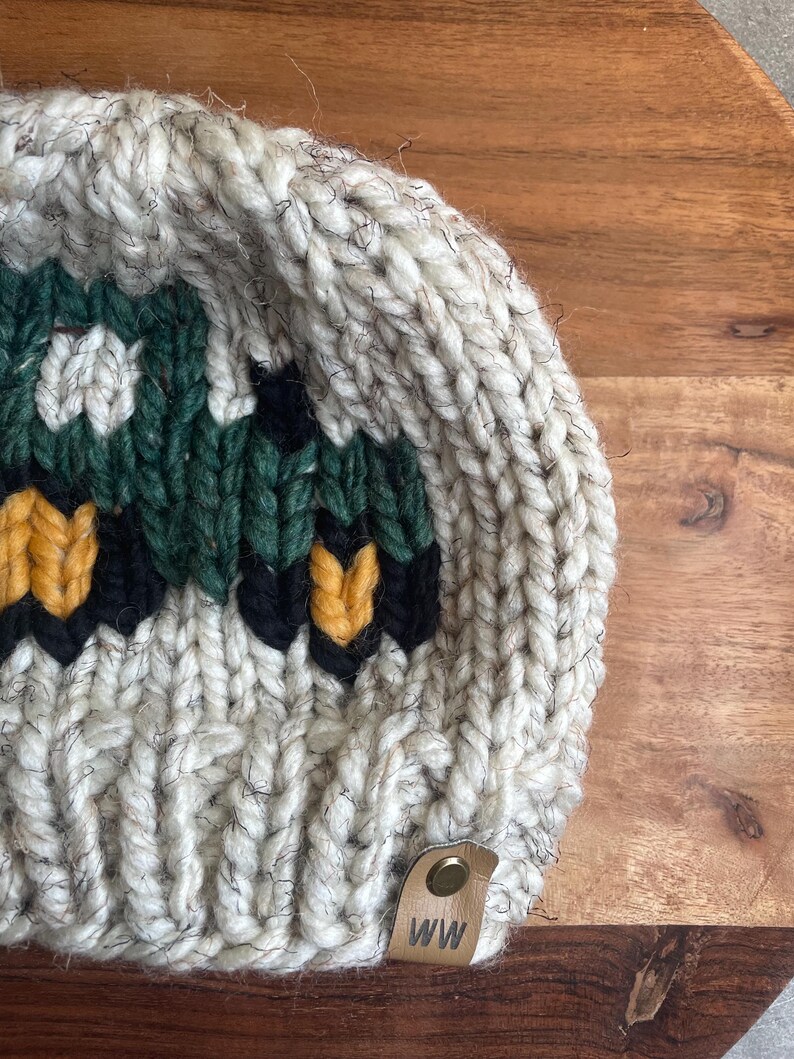 May include: A chunky knit beanie in cream, green, black, and yellow. The hat features a geometric design and a small leather tag with the initials "WW". The beanie is made of thick yarn, perfect for cold weather.