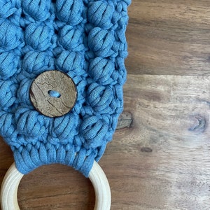 May include: A light blue crocheted fabric piece featuring a textured bobble stitch pattern, secured with a round, dark brown wooden button and attached to a smooth, light-colored circular wooden ring at the bottom edge.
