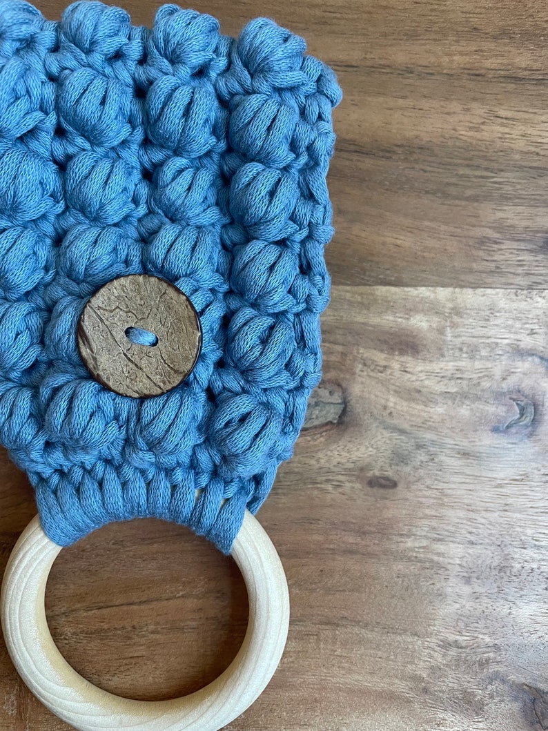 May include: A light blue crocheted fabric piece featuring a textured bobble stitch pattern, secured with a round, dark brown wooden button and attached to a smooth, light-colored circular wooden ring at the bottom edge.