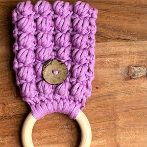 May include: A lavender-colored crocheted fabric piece featuring a textured bobble stitch pattern, secured with a single round brown wooden button, and attached to a smooth, circular light-colored wooden ring at the bottom edge.