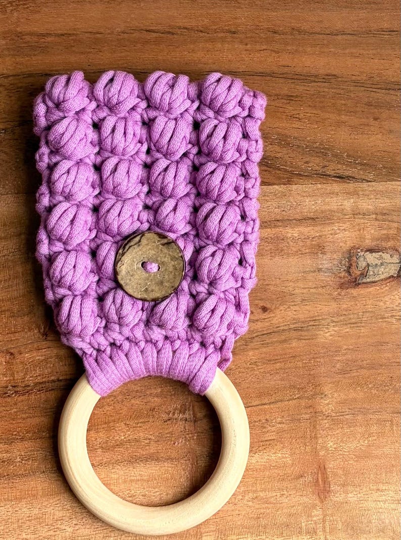 May include: A lavender-colored crocheted fabric piece featuring a textured bobble stitch pattern, secured with a single round brown wooden button, and attached to a smooth, circular light-colored wooden ring at the bottom edge.