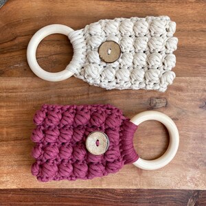 May include: Two macrame towel holders featuring a textured bobble stitch pattern. One holder is cream-colored and the other is deep maroon. Each piece includes a smooth, light-colored wooden ring at one end and a single round wooden button at the center.
