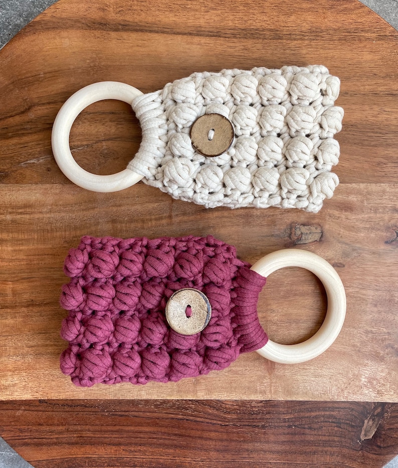 May include: Two macrame towel holders featuring a textured bobble stitch pattern. One holder is cream-colored and the other is deep maroon. Each piece includes a smooth, light-colored wooden ring at one end and a single round wooden button at the center.