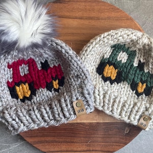 May include: Two knitted beanies with truck designs. One is gray with a red truck, the other is off-white with a green truck. Both have black and yellow accents, and a leather tag with "WW". The gray beanie has a fluffy gray pom-pom.