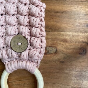May include: A light pink crocheted fabric piece featuring a textured bobble stitch pattern. A circular wooden ring is attached to the bottom edge with a light pink cord. A single round, dark brown wooden button is centered on the crocheted surface.