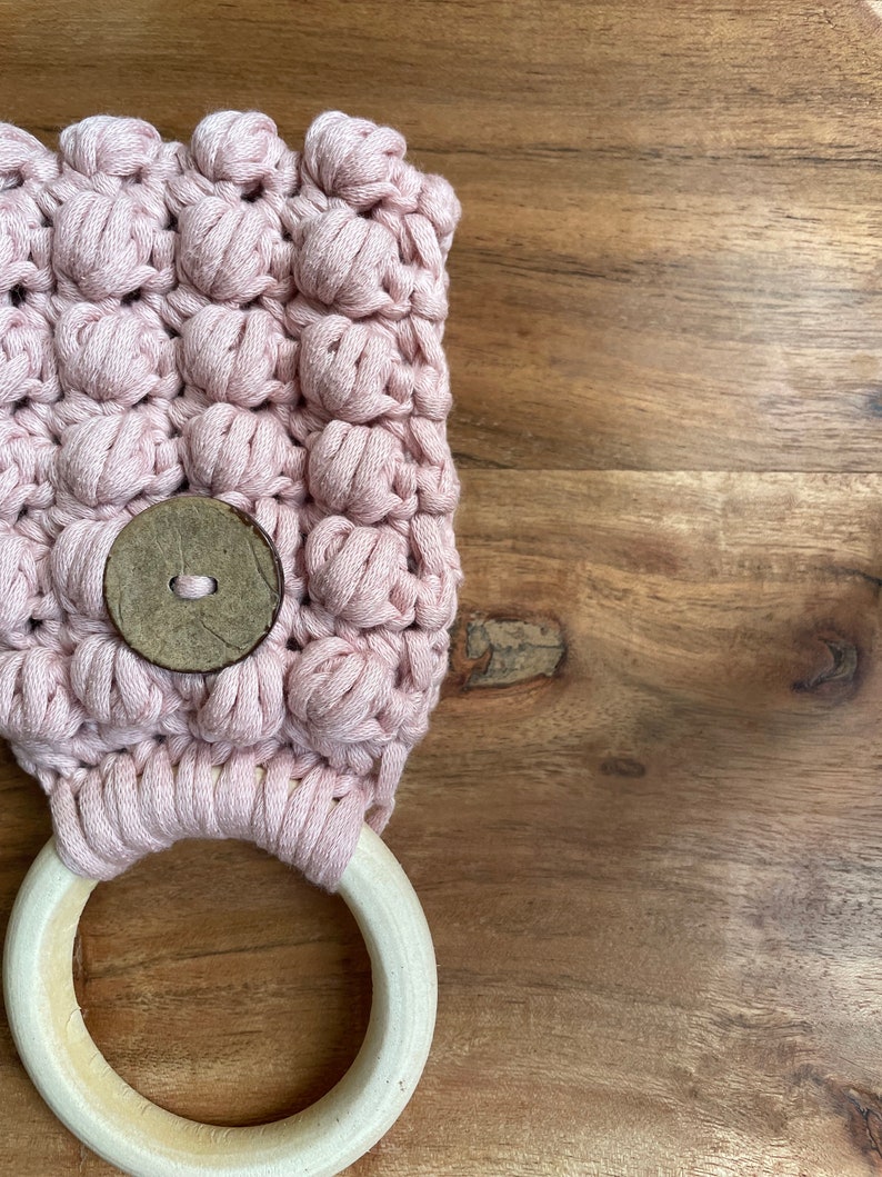 May include: A light pink crocheted fabric piece featuring a textured bobble stitch pattern. A circular wooden ring is attached to the bottom edge with a light pink cord. A single round, dark brown wooden button is centered on the crocheted surface.