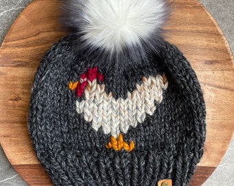 May include: A gray knitted beanie with a white pom pom and a knitted chicken design. The beanie has a brown leather tag with the letters "WW" on it.