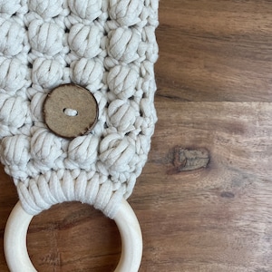 May include: An off-white macrame towel holder featuring a textured bobble stitch pattern, a single round brown wooden button centered on the front, and a smooth, light-colored circular wooden ring attached at the bottom for hanging items.