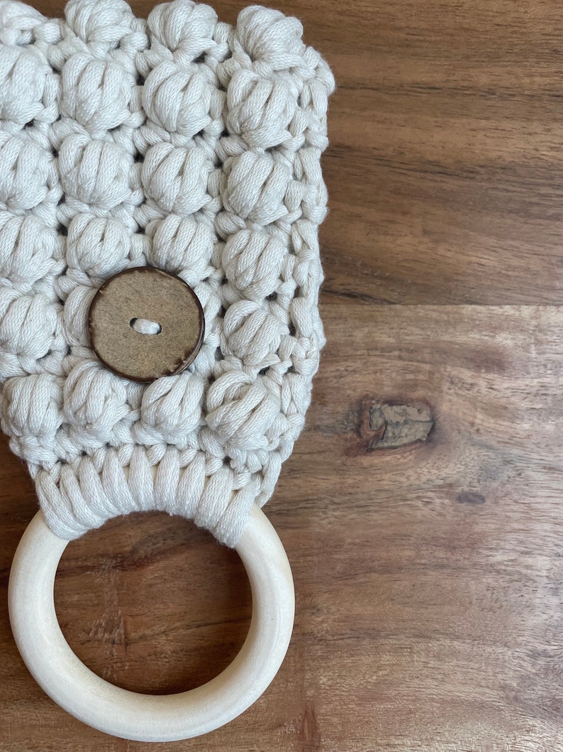 May include: An off-white macrame towel holder featuring a textured bobble stitch pattern, a single round brown wooden button centered on the front, and a smooth, light-colored circular wooden ring attached at the bottom for hanging items.