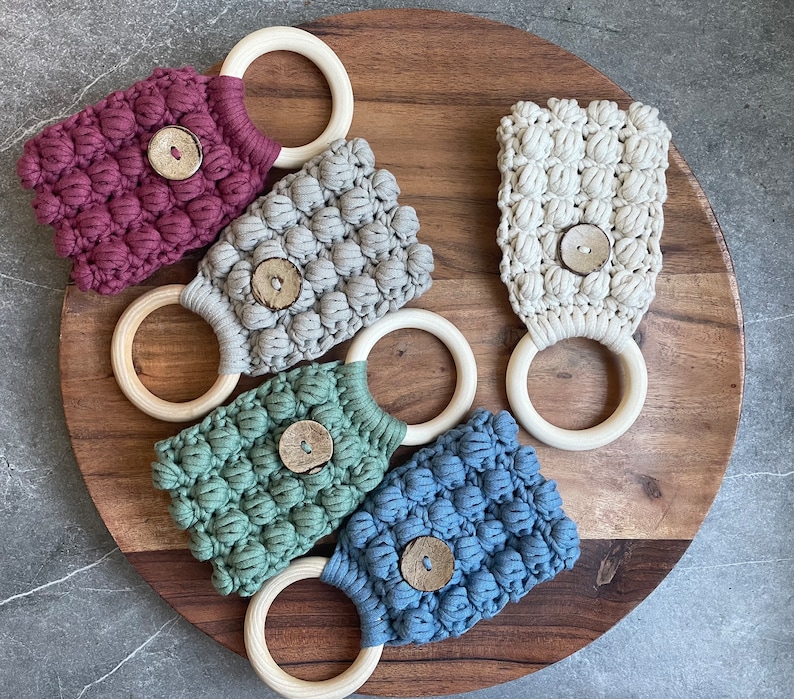May include: Five macrame towel holders in burgundy, light gray, cream, sage green, and dusty blue. Each holder features a textured bobble stitch pattern, a central round wooden button, and a smooth, circular light-colored wooden ring at the top.