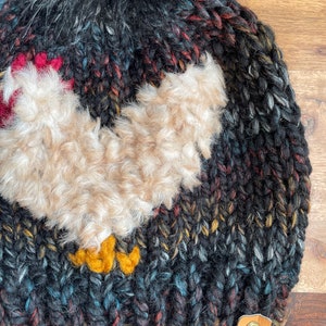 Puede incluir: Gorro de punto negro con un pomp&oacute;n esponjoso y una aplicaci&oacute;n de pollo marr&oacute;n peludo. El gorro tiene un parche de cuero marr&oacute;n con las letras "WW" en &eacute;l.