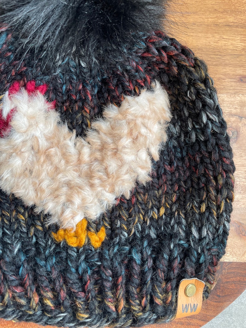 Puede incluir: Gorro de punto negro con un pomp&oacute;n esponjoso y una aplicaci&oacute;n de pollo marr&oacute;n peludo. El gorro tiene un parche de cuero marr&oacute;n con las letras "WW" en &eacute;l.