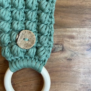 May include: A sage green crocheted fabric piece featuring a textured bobble stitch pattern and a single round, mottled brown button at the center. The fabric is attached to a smooth, light-colored circular wooden ring at the bottom edge.