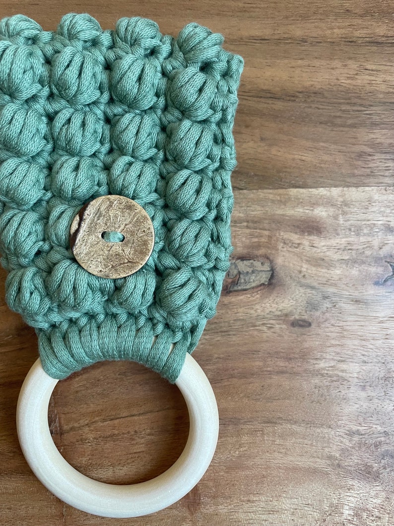 May include: A sage green crocheted fabric piece featuring a textured bobble stitch pattern and a single round, mottled brown button at the center. The fabric is attached to a smooth, light-colored circular wooden ring at the bottom edge.