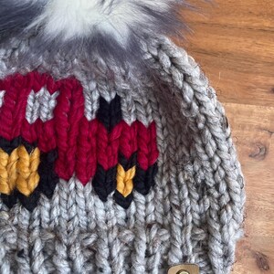 May include: A gray knit beanie with a red, black, and yellow design. The hat features a fluffy, gray and white pom-pom on top and a small leather tag with the initials "WW".