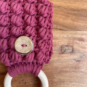 May include: A dark mauve crocheted towel holder featuring a textured bobble stitch pattern and a single round, light-colored wooden button centered on the front. A smooth, unfinished light-colored wooden ring is attached to the bottom of the piece.