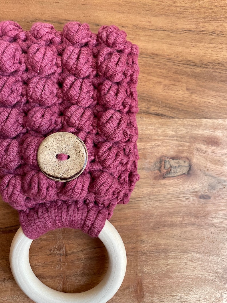 May include: A dark mauve crocheted towel holder featuring a textured bobble stitch pattern and a single round, light-colored wooden button centered on the front. A smooth, unfinished light-colored wooden ring is attached to the bottom of the piece.