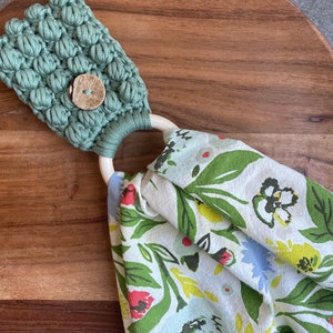 May include: A sage green macrame towel holder featuring a textured bobble stitch pattern and a round wooden button. A light-colored wooden ring connects the macrame top to a white fabric towel printed with a colorful floral pattern of green leaves.