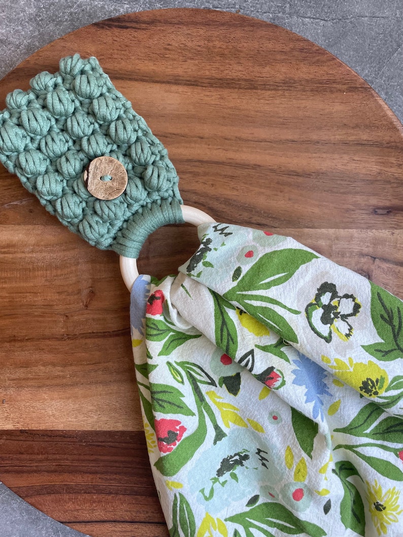 May include: A sage green macrame towel holder featuring a textured bobble stitch pattern and a round wooden button. A light-colored wooden ring connects the macrame top to a white fabric towel printed with a colorful floral pattern of green leaves.