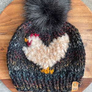 Puede incluir: Gorro de punto negro con un pomp&oacute;n negro esponjoso y una aplicaci&oacute;n de pollo blanco esponjoso. El gorro tiene una etiqueta de cuero marr&oacute;n con las letras "WW" en ella.