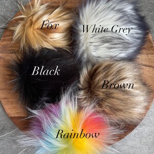 Puede incluir: Cinco pompones de piel sint&eacute;tica en diferentes colores: zorro, gris blanco, negro, marr&oacute;n y arco&iacute;ris. Los pompones est&aacute;n dispuestos en c&iacute;rculo sobre una superficie de madera.