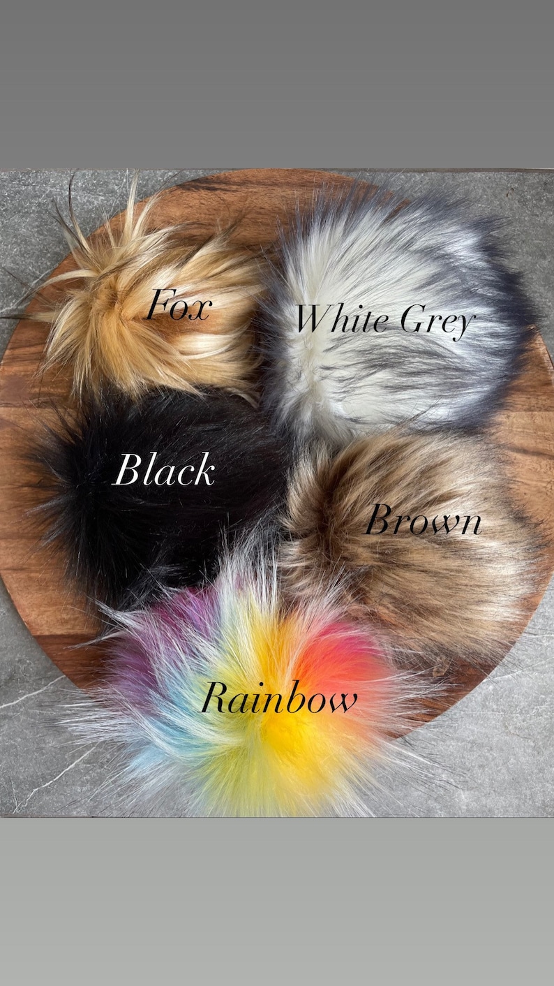 Puede incluir: Cinco pompones de piel sint&eacute;tica en diferentes colores: zorro, gris blanco, negro, marr&oacute;n y arco&iacute;ris. Los pompones est&aacute;n dispuestos en c&iacute;rculo sobre una superficie de madera.