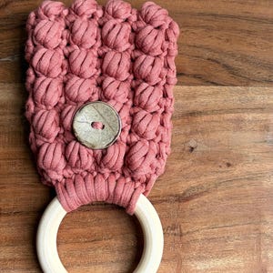May include: A dusty rose macrame towel holder featuring a textured bobble stitch pattern. The top section is secured with a single round, mottled beige button, while the bottom consists of a smooth, light-colored circular wooden ring.