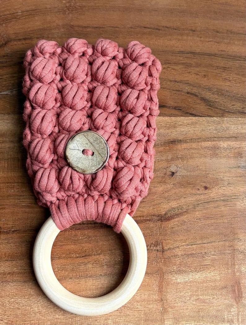 May include: A dusty rose macrame towel holder featuring a textured bobble stitch pattern. The top section is secured with a single round, mottled beige button, while the bottom consists of a smooth, light-colored circular wooden ring.