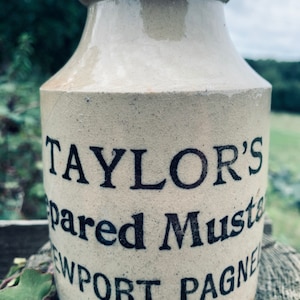 Könnte beinhalten: Vintage Senftopf aus Steinzeug in Beige mit dunkler brauner Kante. Der Topf trägt den schwarzen Text "TAYLOR'S Prepared Mustard NEWPORT PAGNEL". Der Topf ist zylindrisch mit leicht ausgestelltem Oberteil.