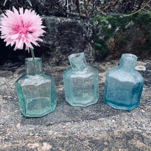 May include: Three small, clear glass bottles with octagonal shapes. The bottles are slightly different shades of blue and green. A pink flower is in the bottle on the left.
