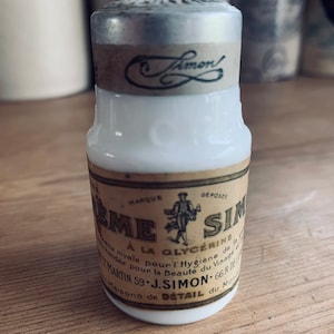 Puede incluir: Una botella de vidrio blanco vintage con tapa plateada y etiqueta marrón. La etiqueta presenta el texto "Crème Simon" y una pequeña ilustración de una persona. La botella es probablemente un producto cosmético.