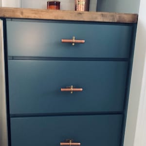 May include: Three drawers with copper drawer pulls on a dark blue cabinet. The cabinet is in a kitchen setting.