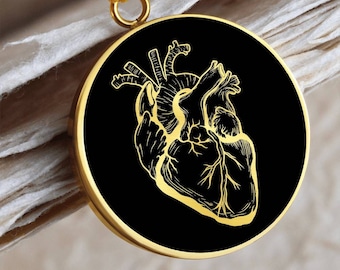 Può includere: Una collana d'oro con un ciondolo circolare nero che presenta un design di cuore anatomico dettagliato.