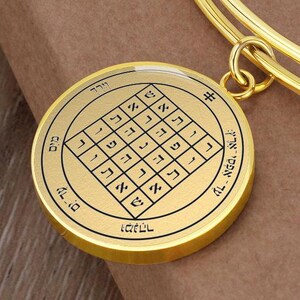Op de afbeelding: Een gouden metalen hanger met een rond ontwerp met een vierkant raster met Hebreeuwse karakters en de woorden "עד־יִם" en "עד־ אַפְסֵי אָרֶץ:".