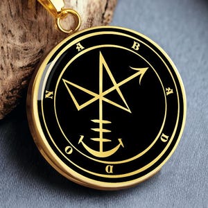 May include: Gold-coloured pendant necklace with a black circular pendant. The pendant features a gold-coloured symbol with the letters A, B, D, and N around the edge. The necklace chain is also gold-coloured.