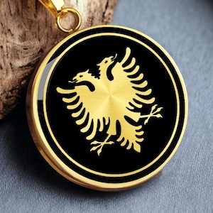 May include: Gold-coloured pendant necklace with a black and gold double-headed eagle emblem. The round pendant hangs from a gold chain. The eagle design is centered on a black background. The necklace rests on a piece of wood.