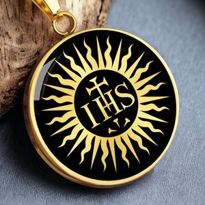 May include: A gold-colored pendant necklace with a circular pendant featuring a black background and a gold-colored sunburst design. The center of the pendant displays a cross above the letters IHS. The necklace is displayed on a wooden surface.
