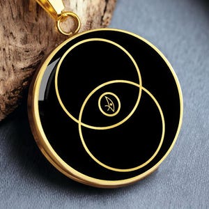 May include: Gold-toned necklace with a circular pendant. The pendant features a black background with two overlapping gold circles and a small symbol. The necklace chain is also gold.
