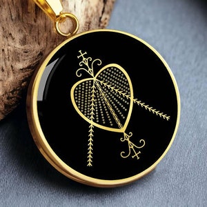 Könnte beinhalten: Eine goldene Kettenkette mit einem runden Anhänger mit schwarzem Emaille-Hintergrund und einem goldenen Herzdesign mit einem stilisierten Pfeilmotiv.