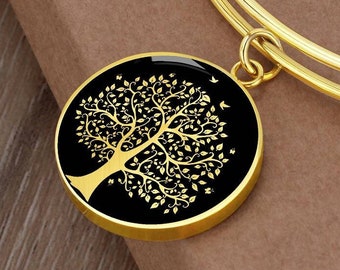 May include: A gold-toned bangle bracelet with a round charm featuring a black background and a gold tree of life design. The charm has small birds flying around the tree.