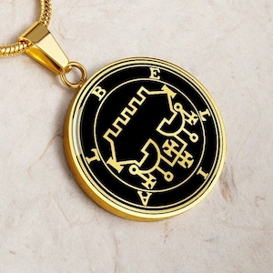 Puede incluir: Un collar de cadena dorada con un colgante negro y dorado que presenta un sello con las letras 'BEITH' y 'EVIL' alrededor del perímetro.
