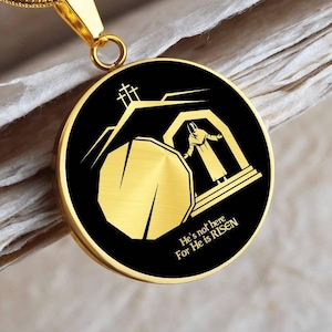 May include: Gold necklace with a round pendant featuring a black and gold illustration of a tomb with a stone rolled away and a figure standing outside. The pendant has the text "He's not here For He is RISEN".