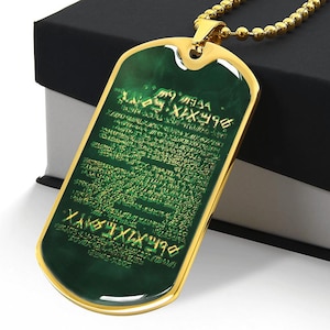May include: A gold-coloured dog tag pendant with a green centre and gold text. The pendant hangs from a gold ball chain. The dog tag is rectangular with rounded corners. The pendant is resting on a black box.